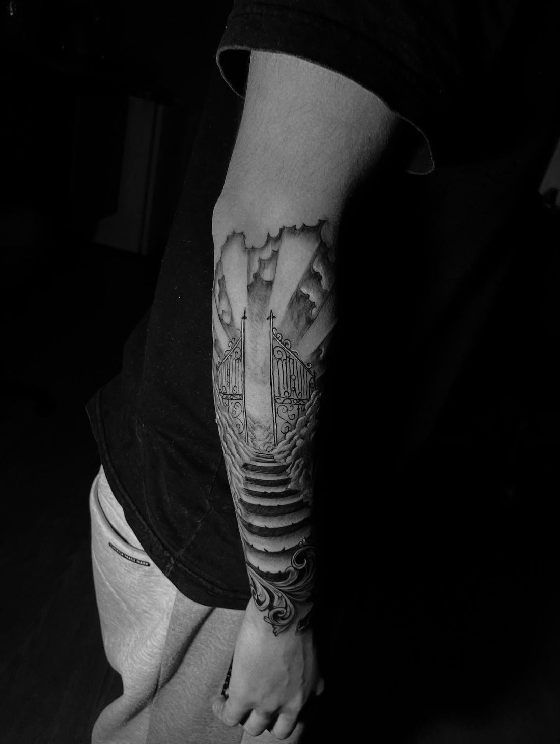 Antebraço tatuado em preto e cinza com portão aberto entre nuvens, raios de luz e escada