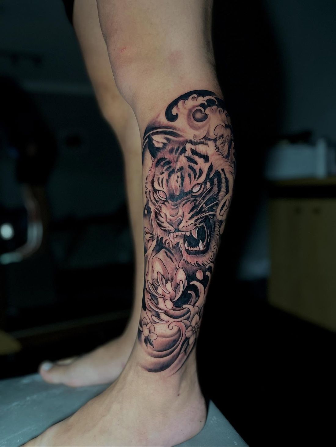 Tatuagem na panturrilha em preto e cinza com tigre rugindo entre ondas e flores de cerejeira